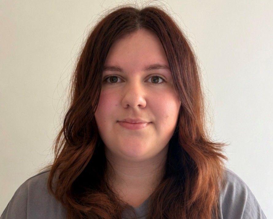 Faye, a woman with long brunette hair in a grey T-shirt, looks at the camera, standing in front of a plain white background