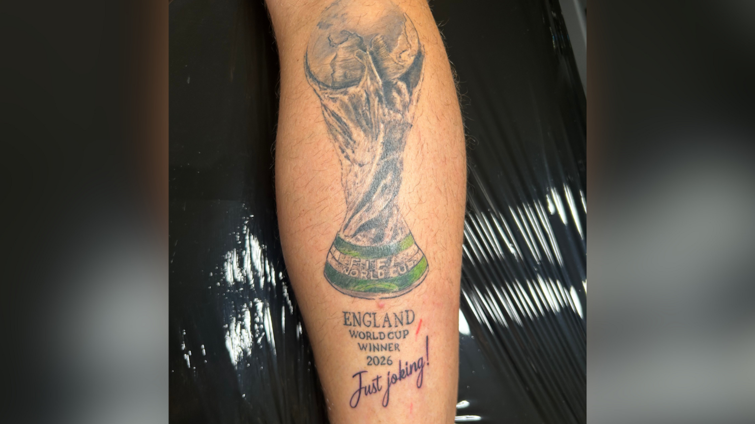 Large tattoo on a man's leg. It is a trophy with a caption reading: 'England World Cup winners 2026.' with a line underneath reading: 'Just joking!'.
