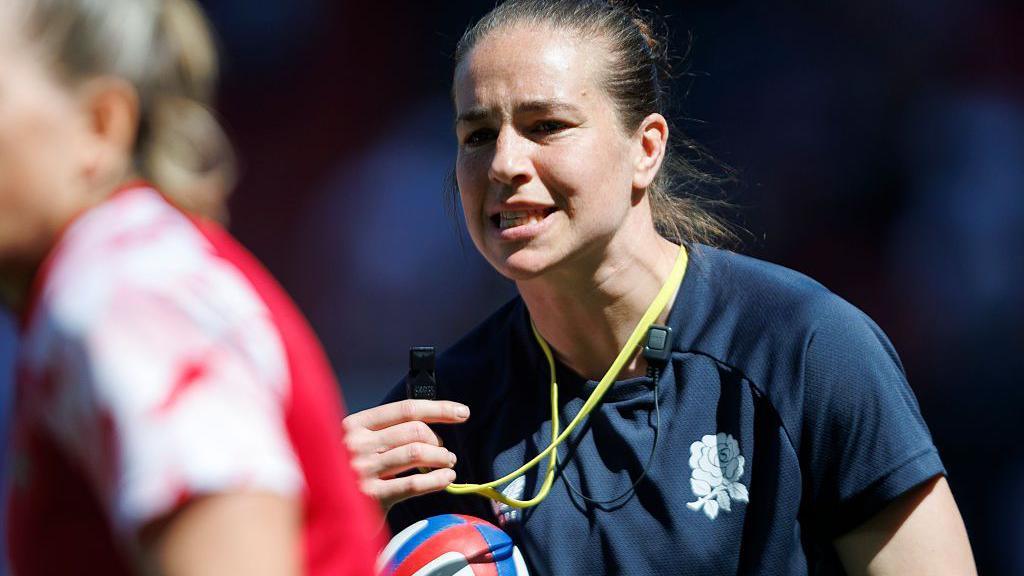 Emily Scarratt brings a whistle to her mouth while holding a rugby ball