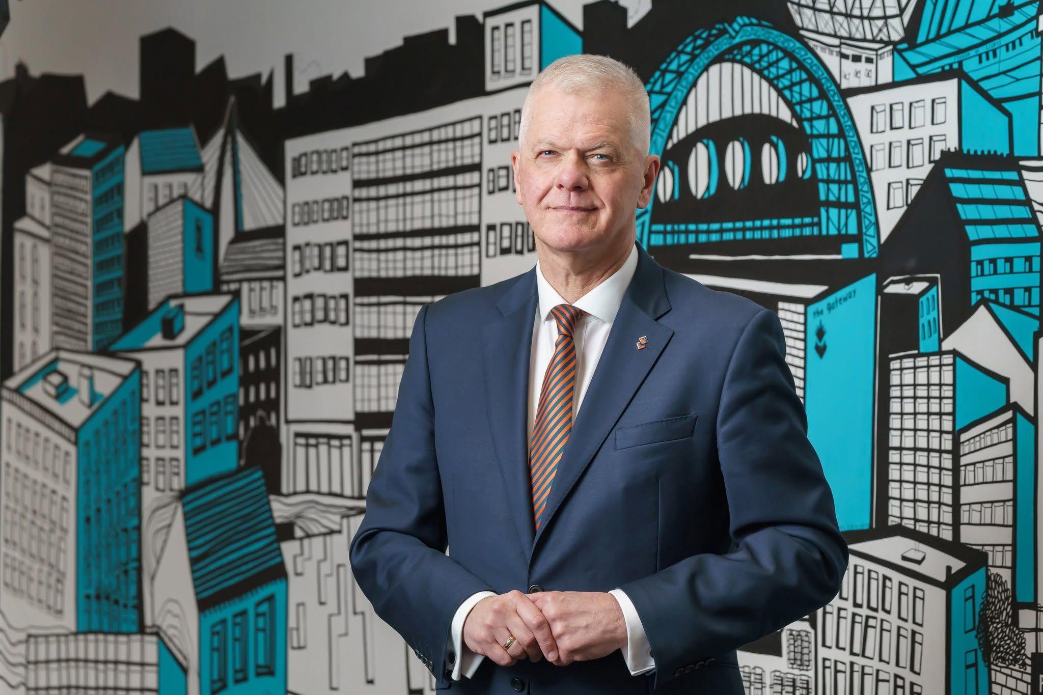 A man with close-cropped white hair, wearing a suit, holds his hands in front of an illustrated mural of Sunderland