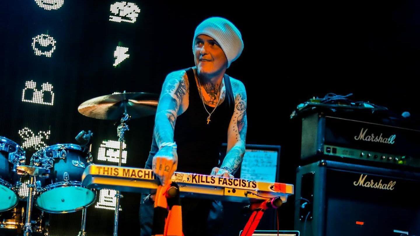 Derry Brownson, the keyboardist of EMF, smiles as he performs on stage. He is wearing a grey beanie, a black tank top and has arms covered in tattoos.