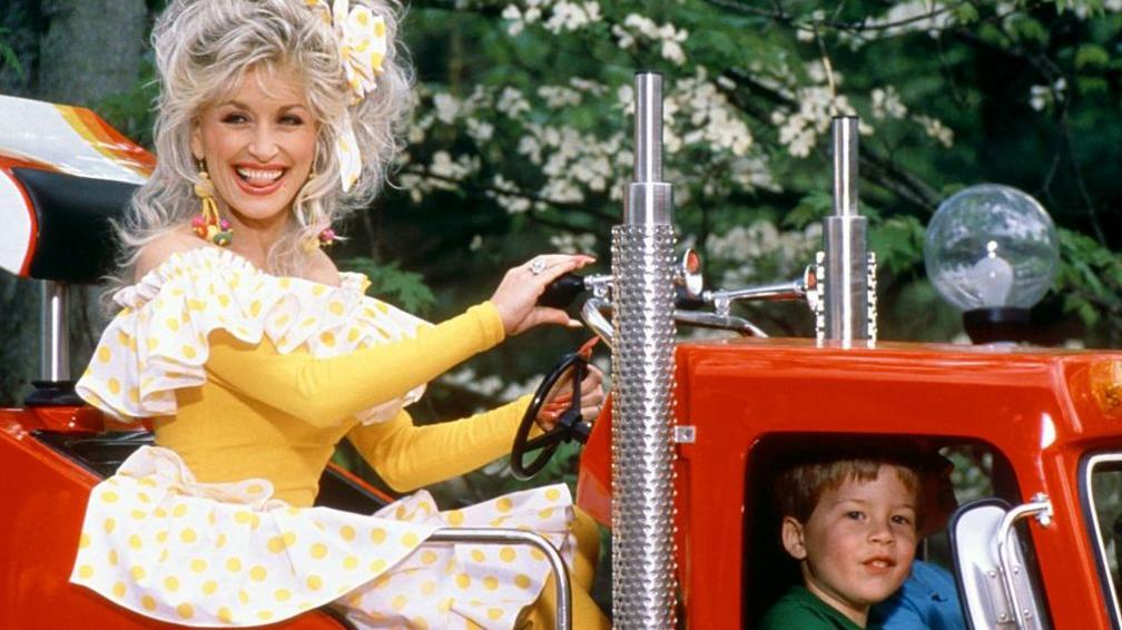 Dolly Parton is pictured riding on a truck at her famous Dollywood theme park in Tennessee in 1977, she's in a yellow and white dresss