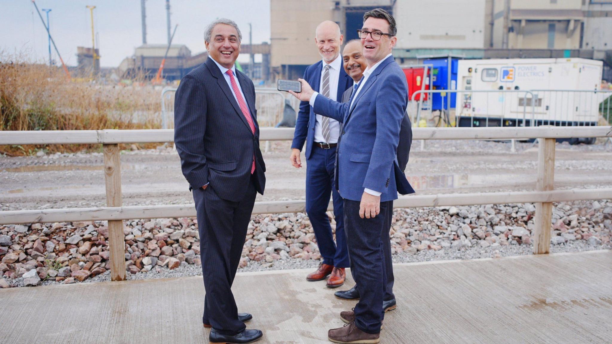 Andy Burnham, wearing a blue jacket and dark trousers, stands with three other men while he takes a selfie outside an industrial site. They are all smiling and laughing.