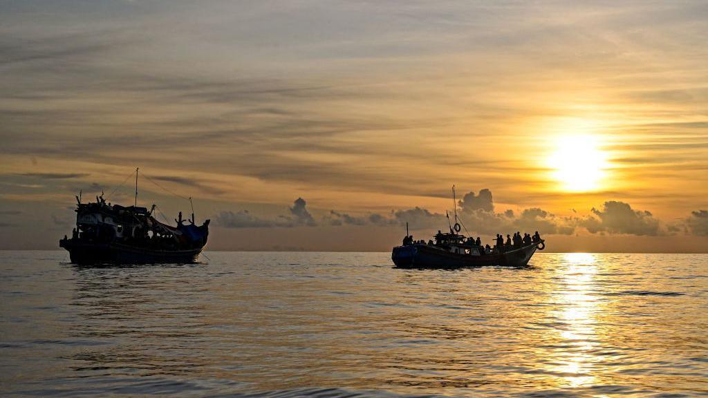 Two boats carrying lots of people float in a sea against a setting sun