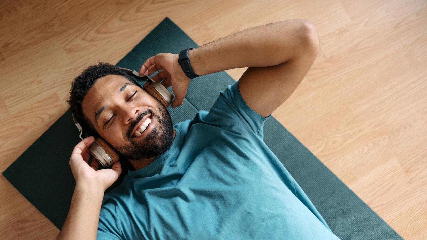 Man wearing blue t-shirt, lying on a dark green yoga mat on a wooden floor, with over-ear headphones on. He's smiling and has his eyes closed.