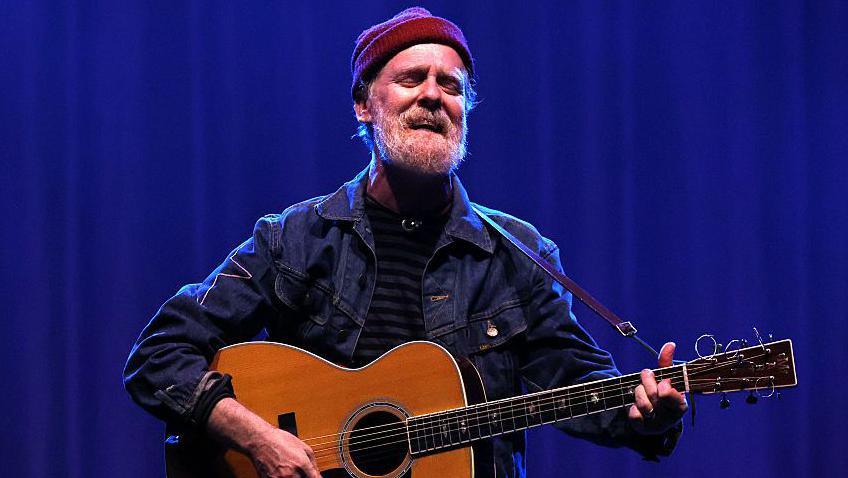 Glen Hansard sings while holding an acoustic guitar. He has a grey beard and is wearing a red wool hat and denim jacket. He is standing in front of a dark blue curtain.