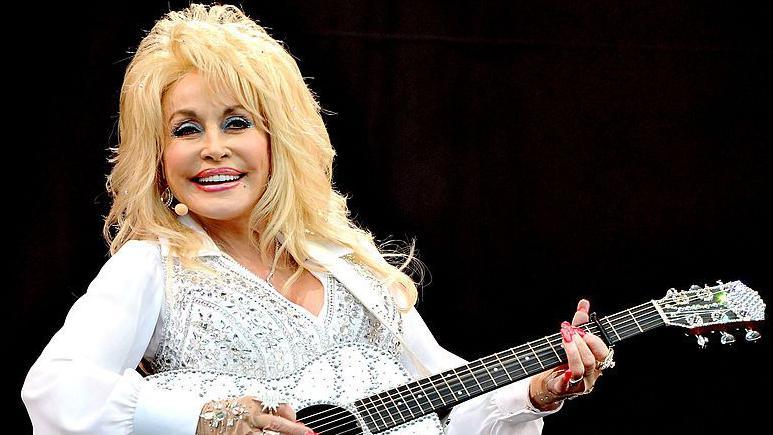 doll parton in a white rhinestone top and a matching acoustic guitar. she has blonde hair and full make up and a big smile as she stands on stage in front a dark background