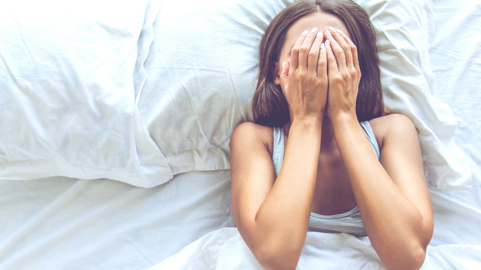 A young woman lies in bed trying to get to sleep. She covers her face with her hands.