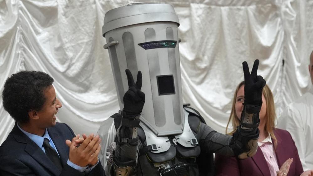 A man wearing a costume which involves a grey bin covering his head is stood in the middle. He is doing a 'V' symbol with both hands. A man and a woman stood either side of Count Binface are looking and smiling at him.