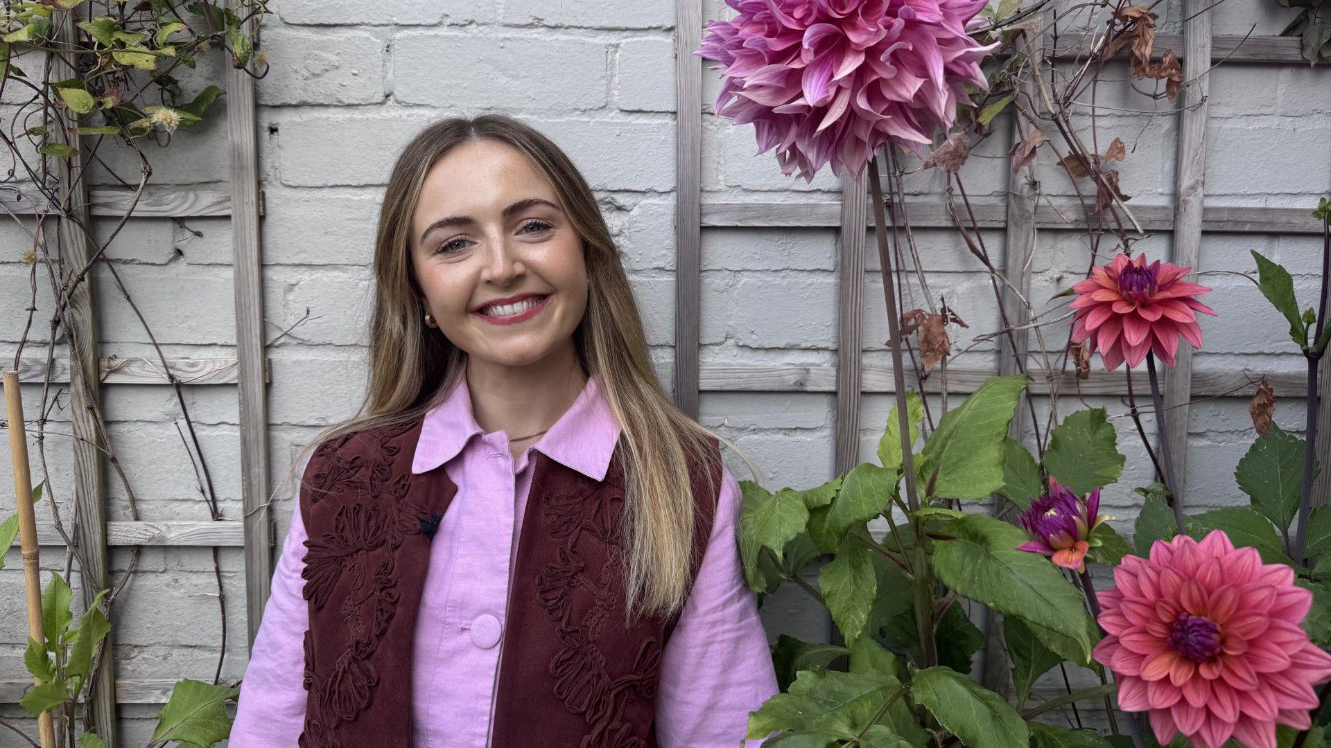 Hannah is a woman in her early 30s. She has long blonde hair and is smiling at the camera. She's pictured against a white brick wall and has tall dahlias growing either side of her. She is wearing a pink collared shirt with a dark red waistcoat.