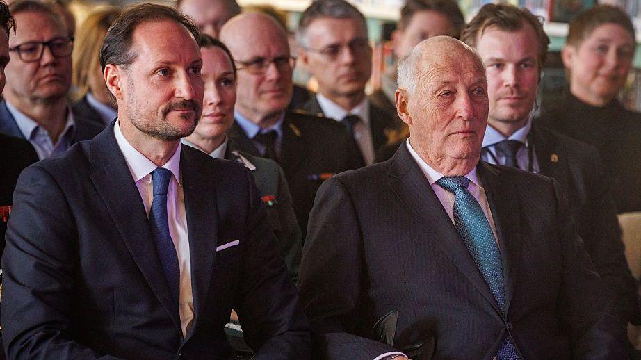 Two men in suits sit sombrely listening in an audience - the older man sits on the right