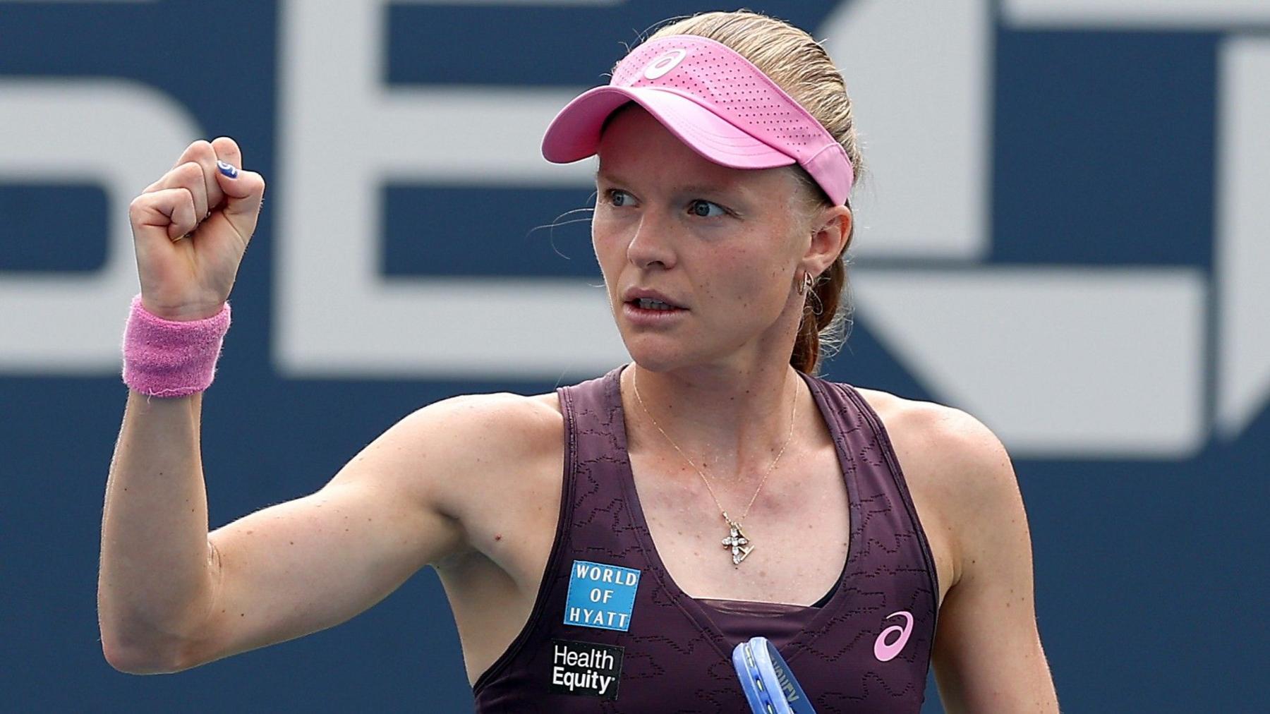 Harriet Dart, wearing a purple dress and pink visor, holds her racquet in her left hand and raises a clenched right fist