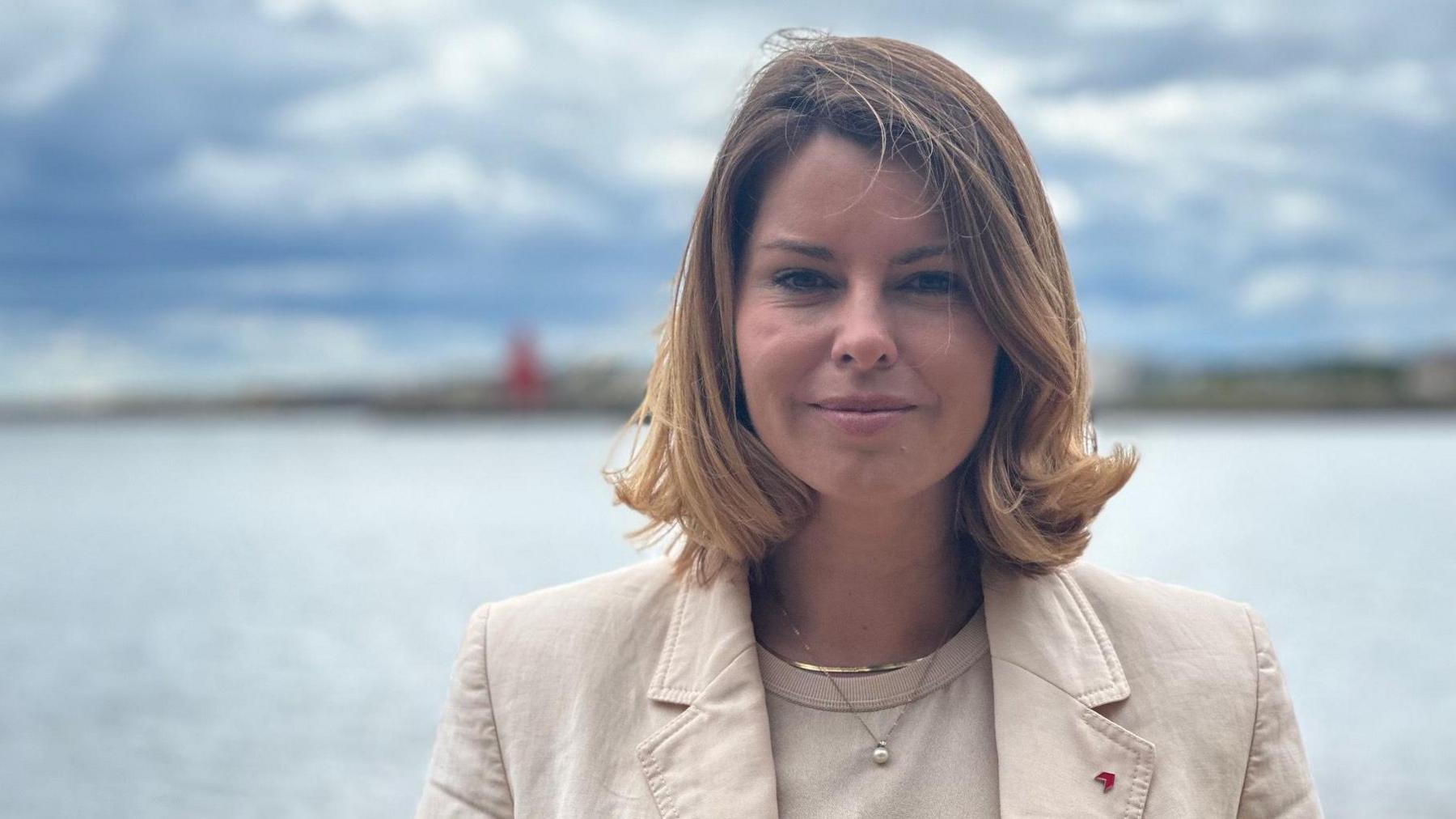 Kim McGuinness is standing with her back to the sea on a beach. She has blonde, shoulder length hair, is half smiling, and is wearing a cream top and cream jacket. In the background, out of focus, is a lighthouse on a pier jutting out into the water.