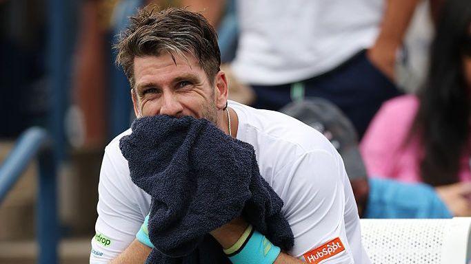 Cameron Norrie, with dark hair and wearing a white t-shirt, covers the lower half of his face with a dark blue towel