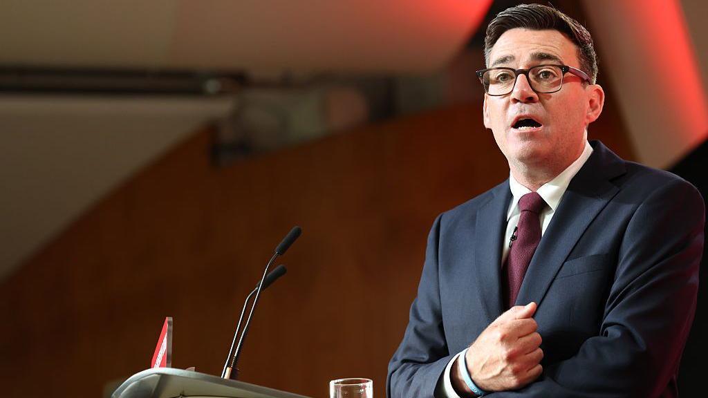 Andy Burnham stood at a lectern delivering his first speech as the leader of the Labour Party. His right hand is clenched in a fist and pulled towards his chest as he speaks. He is wearing glassed, a black suit, white shirt, and burgundy tie.