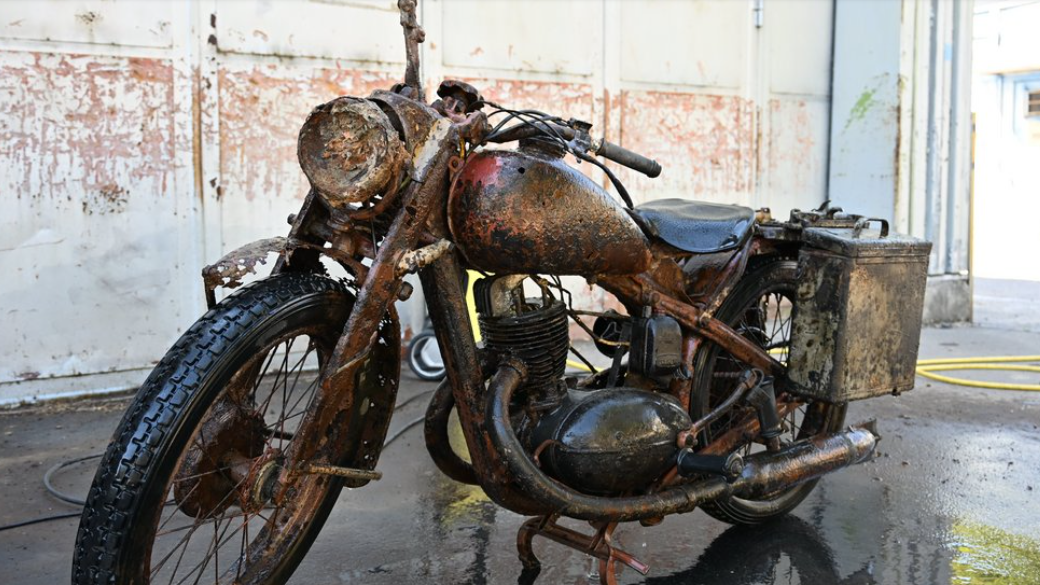 A rusted but largely well-preserved World War Two-era Wehrmacht motorcycle pictured parked in a building resembling a warehouse.
