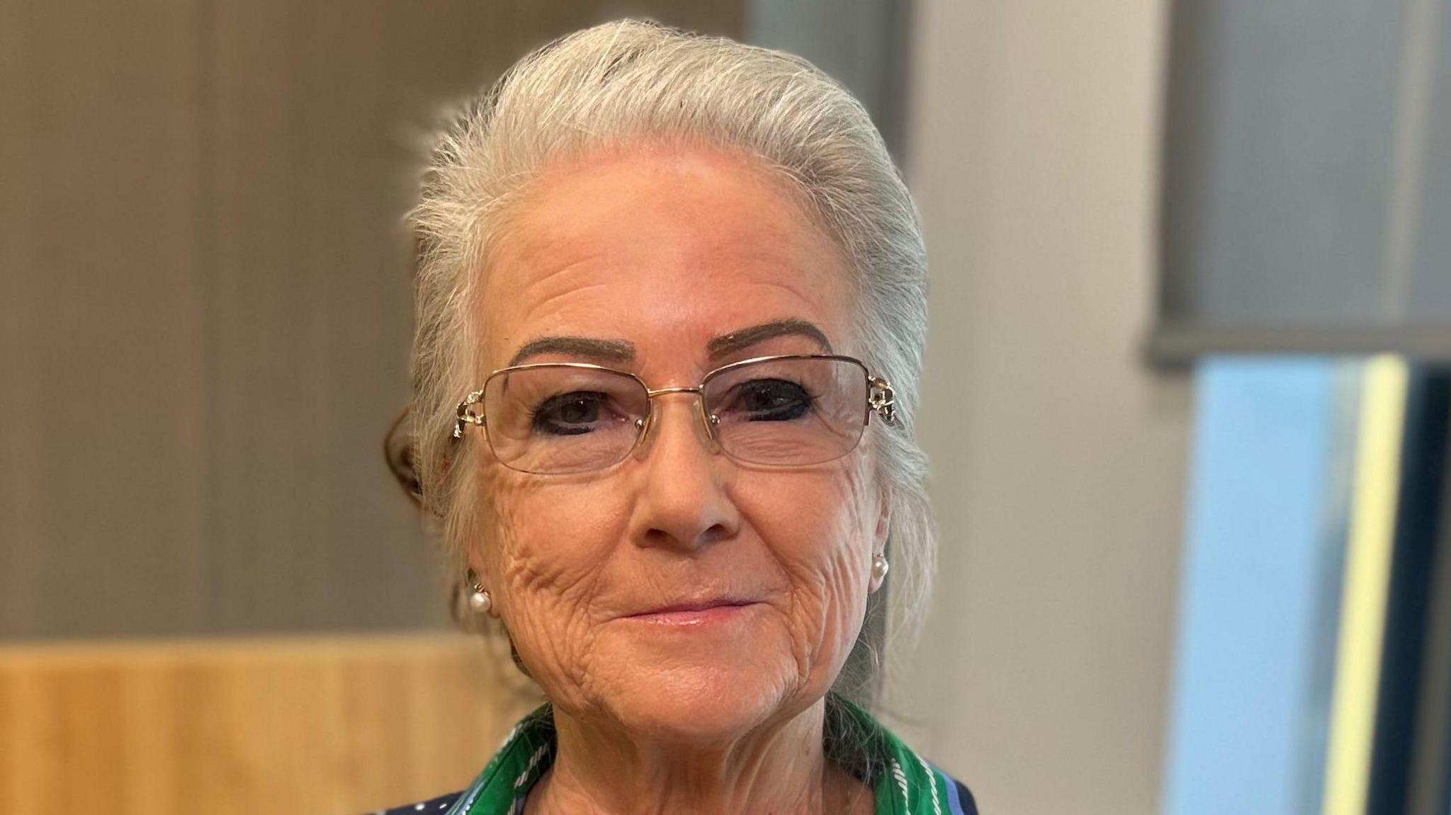 A woman with silver hair looks at the camera. She is wearing small silver-framed glasses