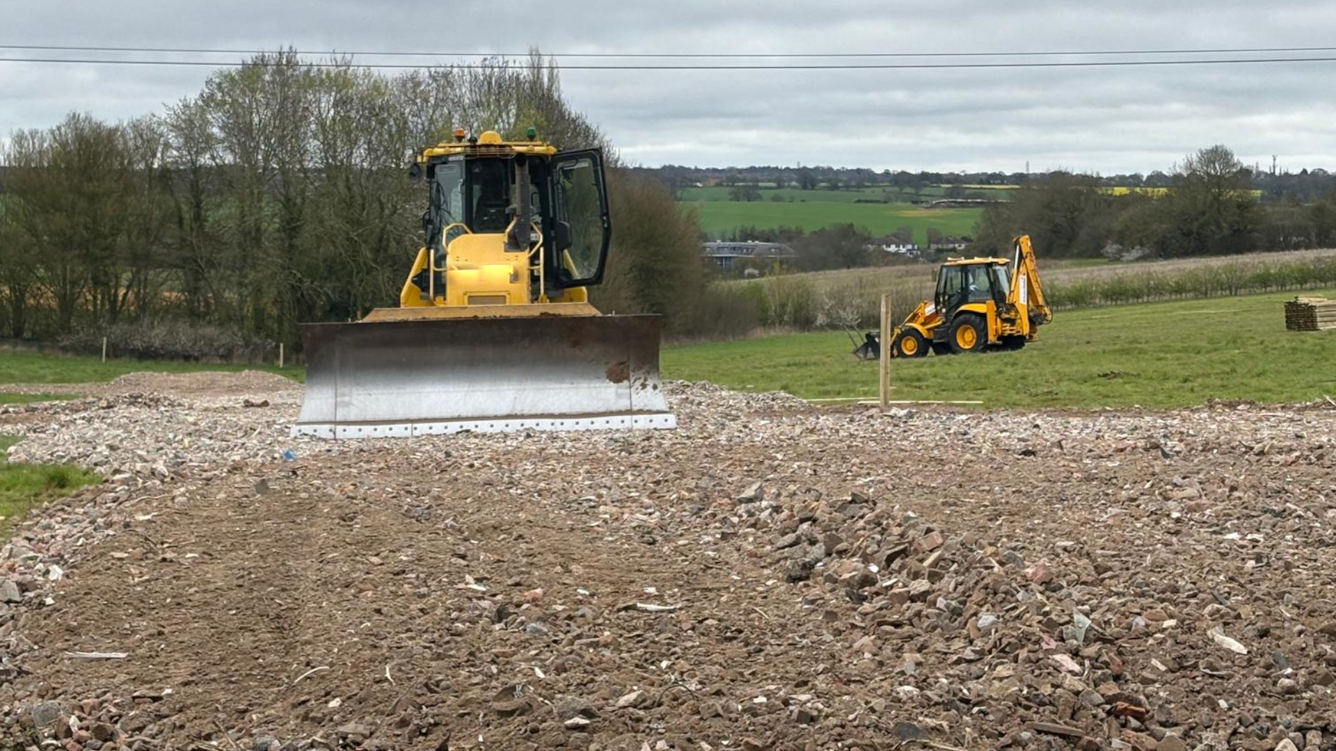A yellow bulldozer faces the camera with its silver scraper at the front. It sits on a large area of soil that is amid a grassy field, and the soil appears to have been recently turned over. There are trees and fields in the background.