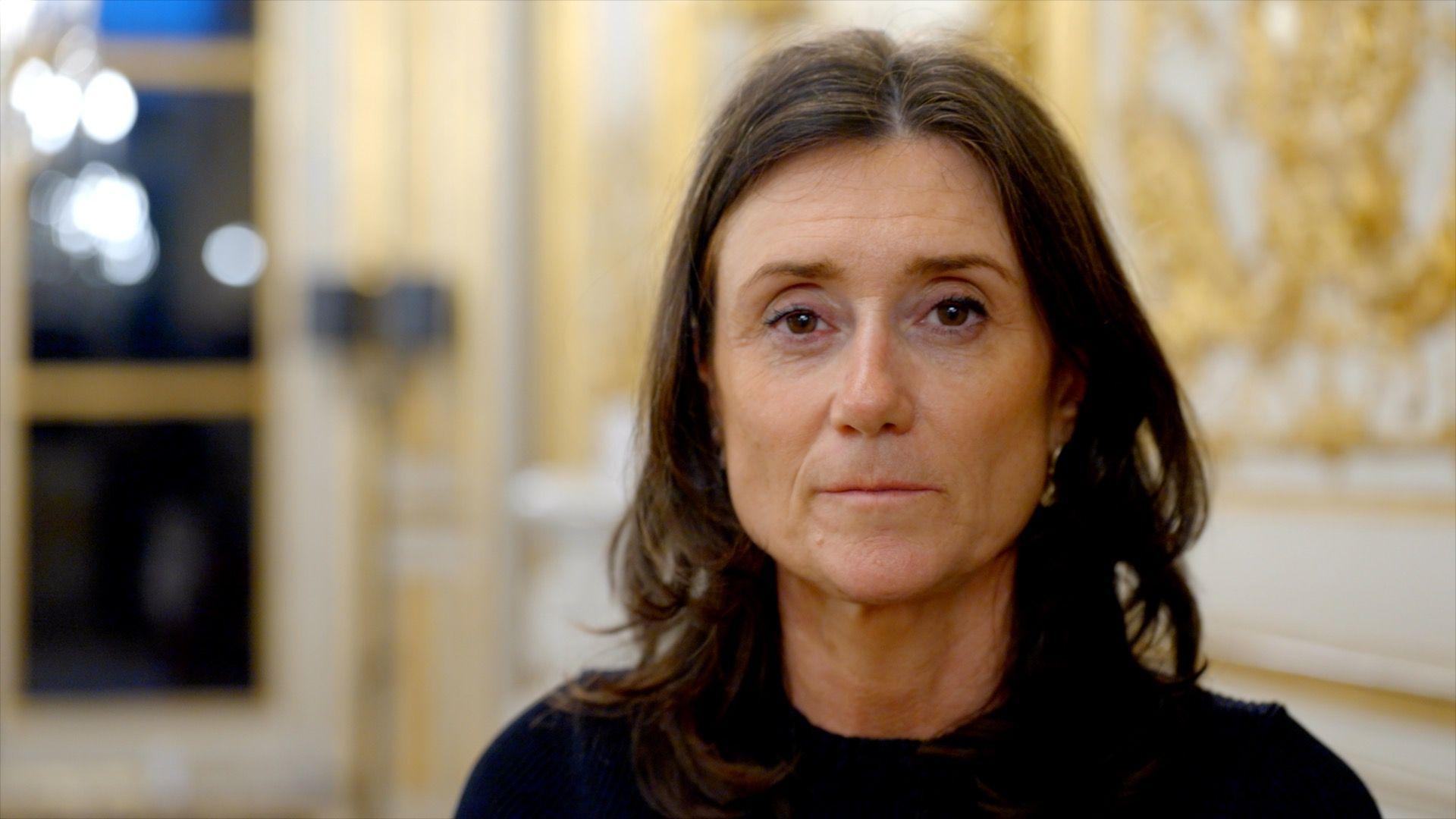 Sandrine Josso stares at the camera. She has mid-length dark hair and is wearing a black top. She's standing in front of a wall with gold leaf on it.