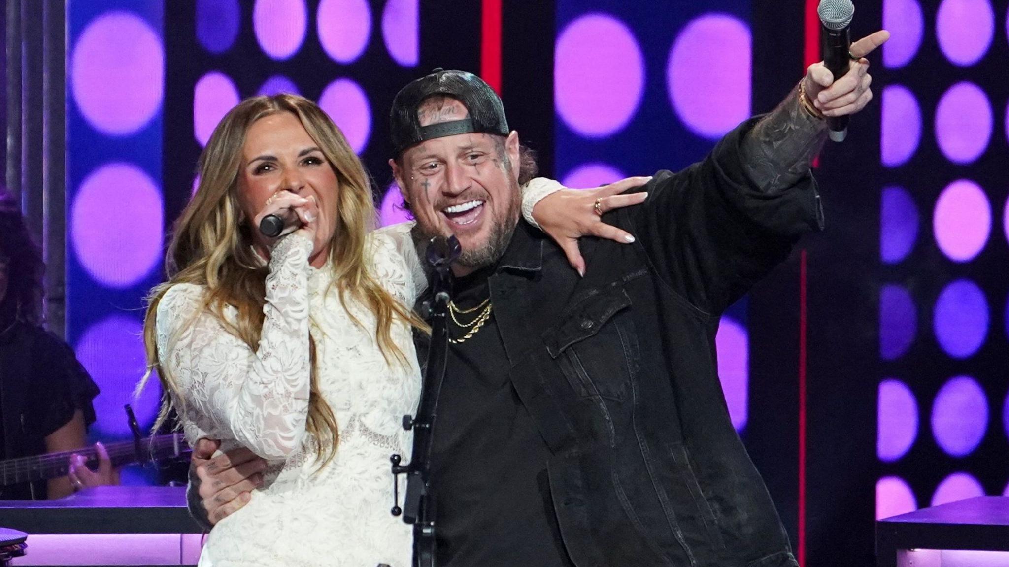 Jelly Roll in black denim jacket and backwards black baseball cap, with one arm around Carly Pearce and the other holding a microphone and pointing in the air. Carly Pearce is weating a white lace dress and singing into her handheld microphone.