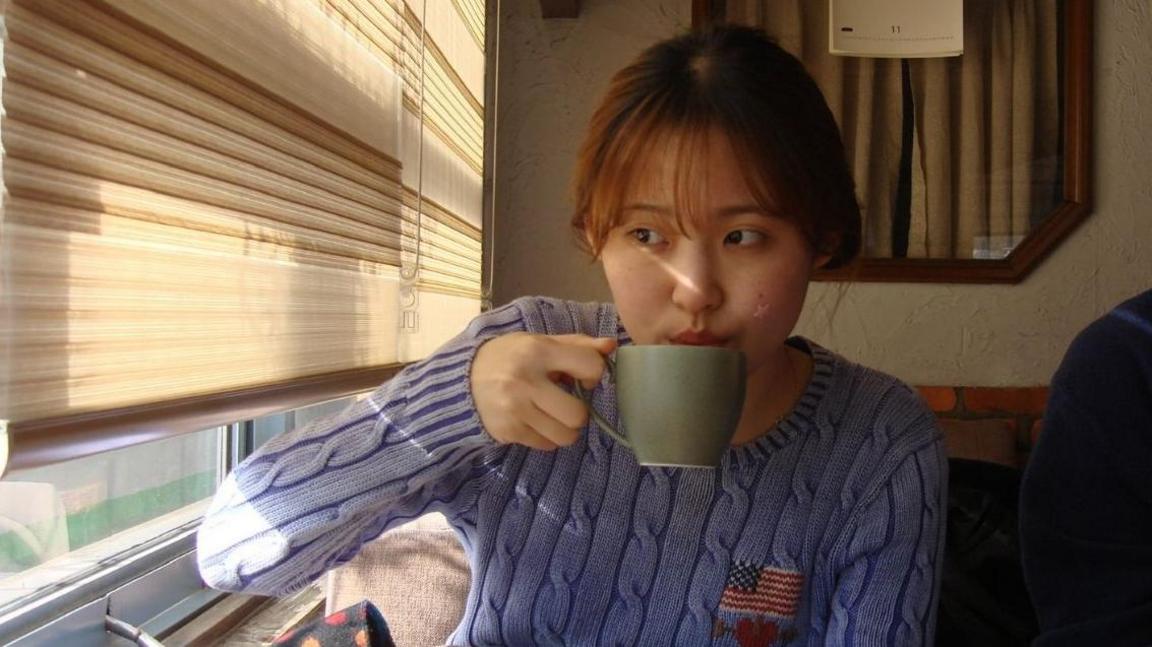 Soomin Yi sipping on a cup of tea while glancing towards a window to her right