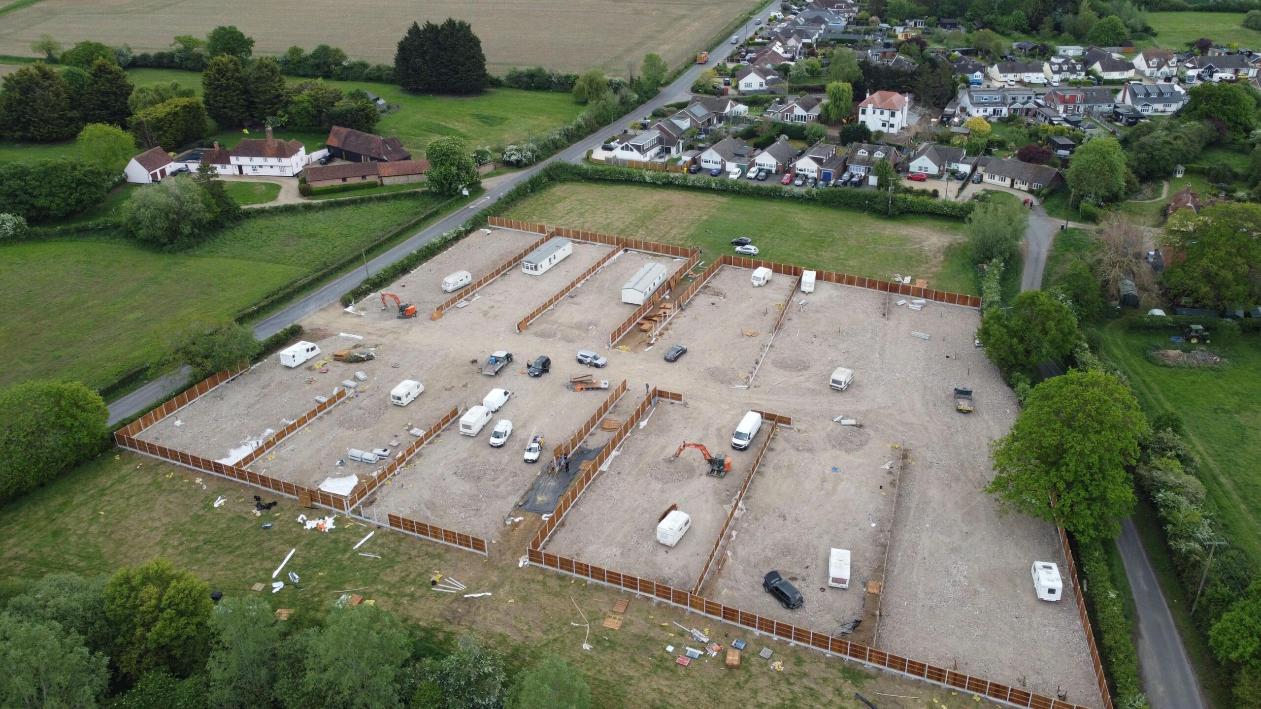 A drone image of the developed site. It is a field that has been largely concreted over and has several caravans, both static and mobile, on it. Fencing is being installed around the perimeter of the site, which is flanked by residential housing and other fields.