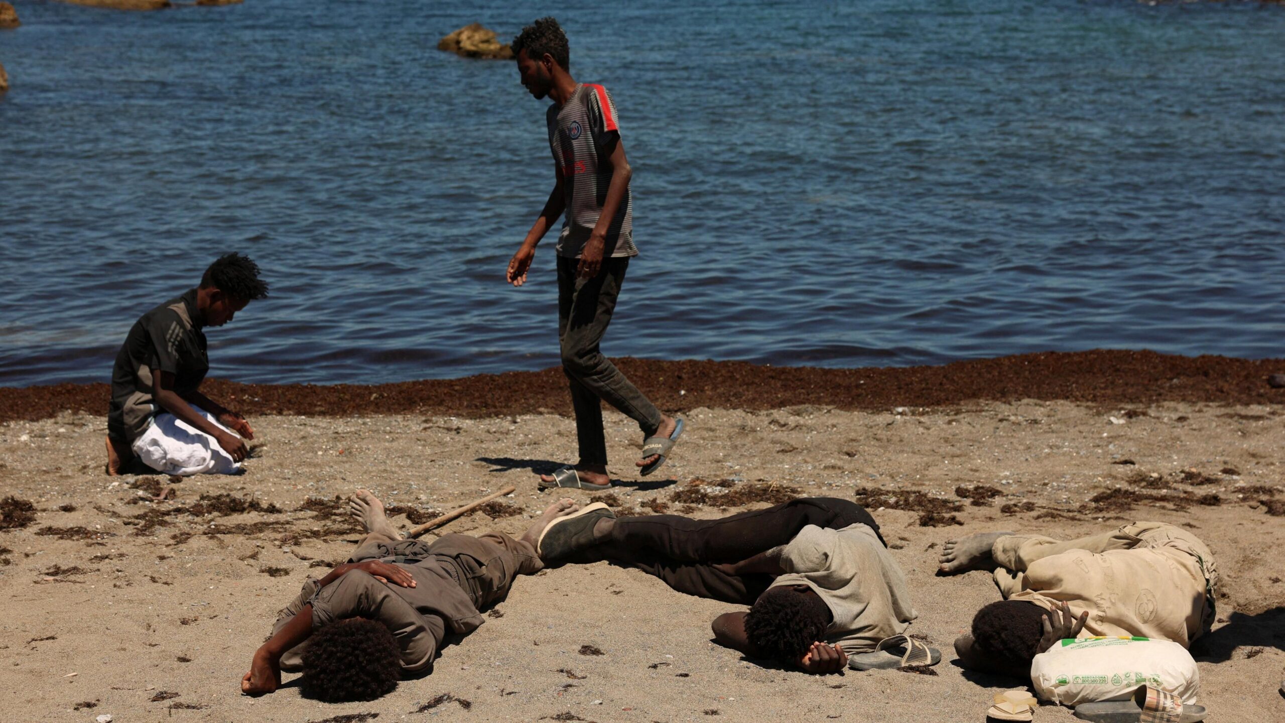 Three men sleep on a beach while another prays in front of the sea in Ceuta