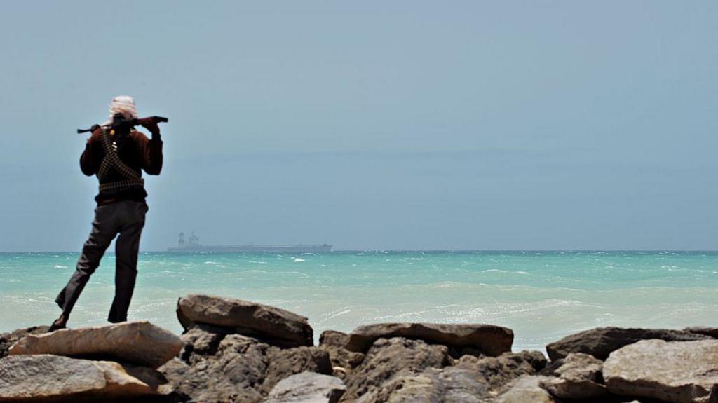 A man - with his back to the camera - stands on some rocks to the left of the picture. Holding a gun across his shoulders, he is looking out to sea where a tanker is in the far distance.