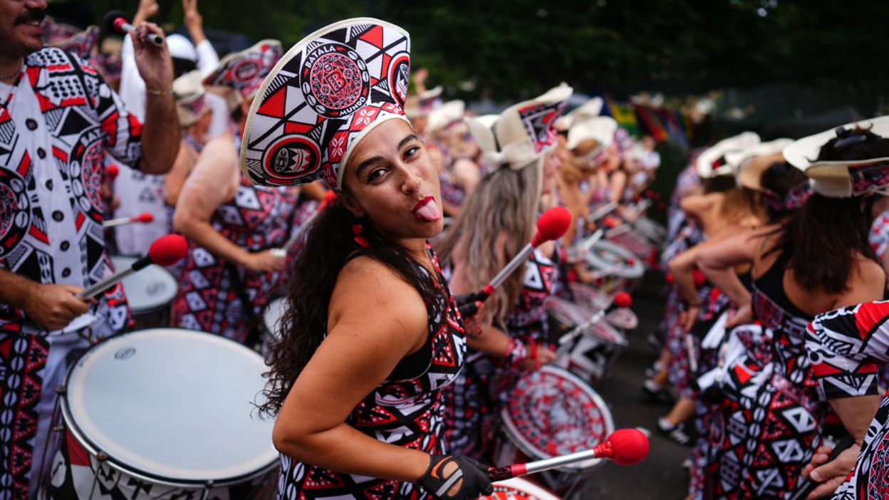 A woman in a black, red and white costume marches along the parade as part of a performing band. She faces the camera and sticks out her tongue.