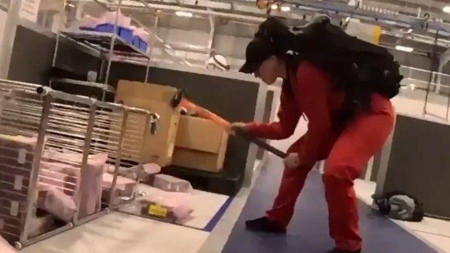 A person wearing a red jumpsuit and black cap, using a sledgehammer to damage equipment inside the factory.