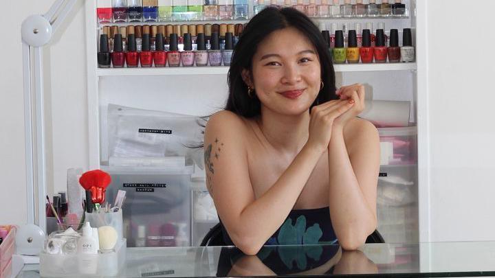Nail artist Jess Young sits behind a table and smiles at the camera. On the table there are nail tools and behind her are nail polish bottles in a range of colours.