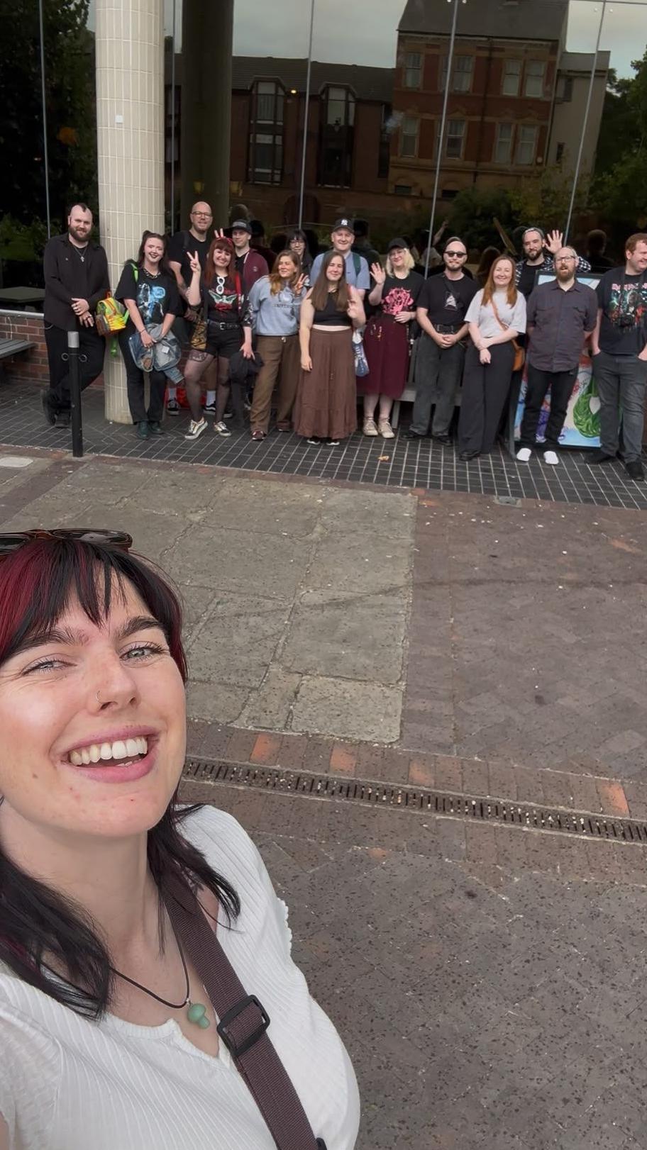 A woman with red and black hair takes a selfie with a large groups of people in the background in front of a museum.