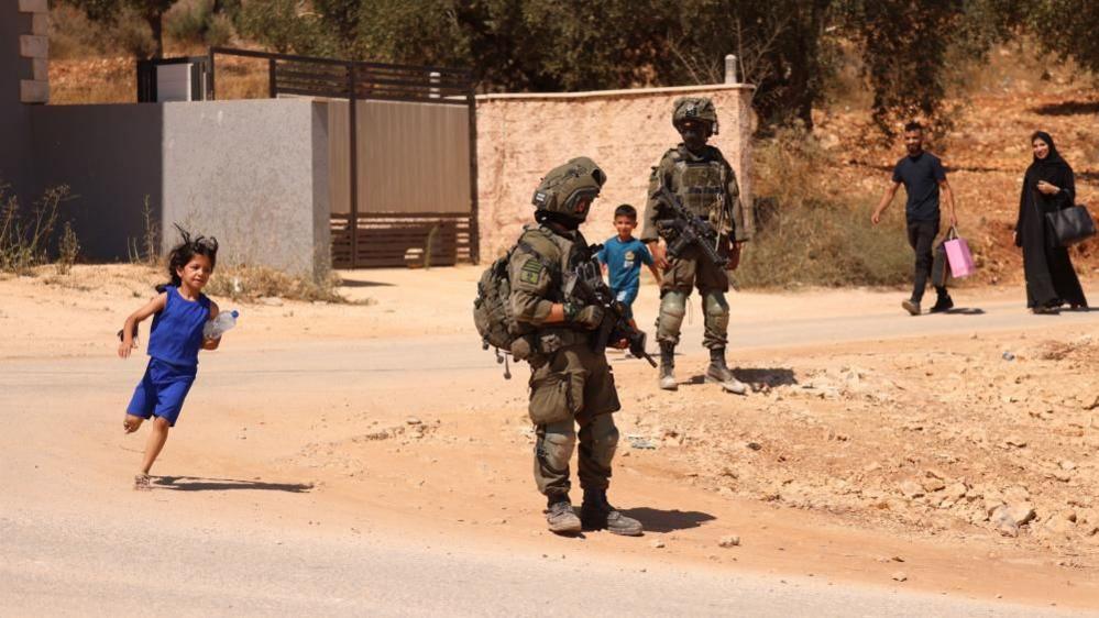 Two soldiers armed and in full uniform stand on the side of a dusty road where a man wearing black clothes and carrying two shopping bags - one dark, one pink - and a woman wearing a black dress and head cover and carrying a black bag are walking. Two children - a boy and a girl - appear to be running, the girl wearing blue shorts and blue top and carrying a large bottle of water under her left arm.