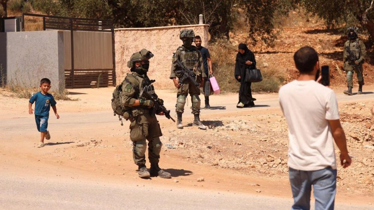 A wide shot showing three IDF soldiers in military gear wearing face coverings and carrying rifles walking along a dusty road. Four West Bank residents including two men, one woman and one boy can be seen walking around the soldiers. One West Bank resident can be seen holding a smartphone up to his face to film or take a photo.