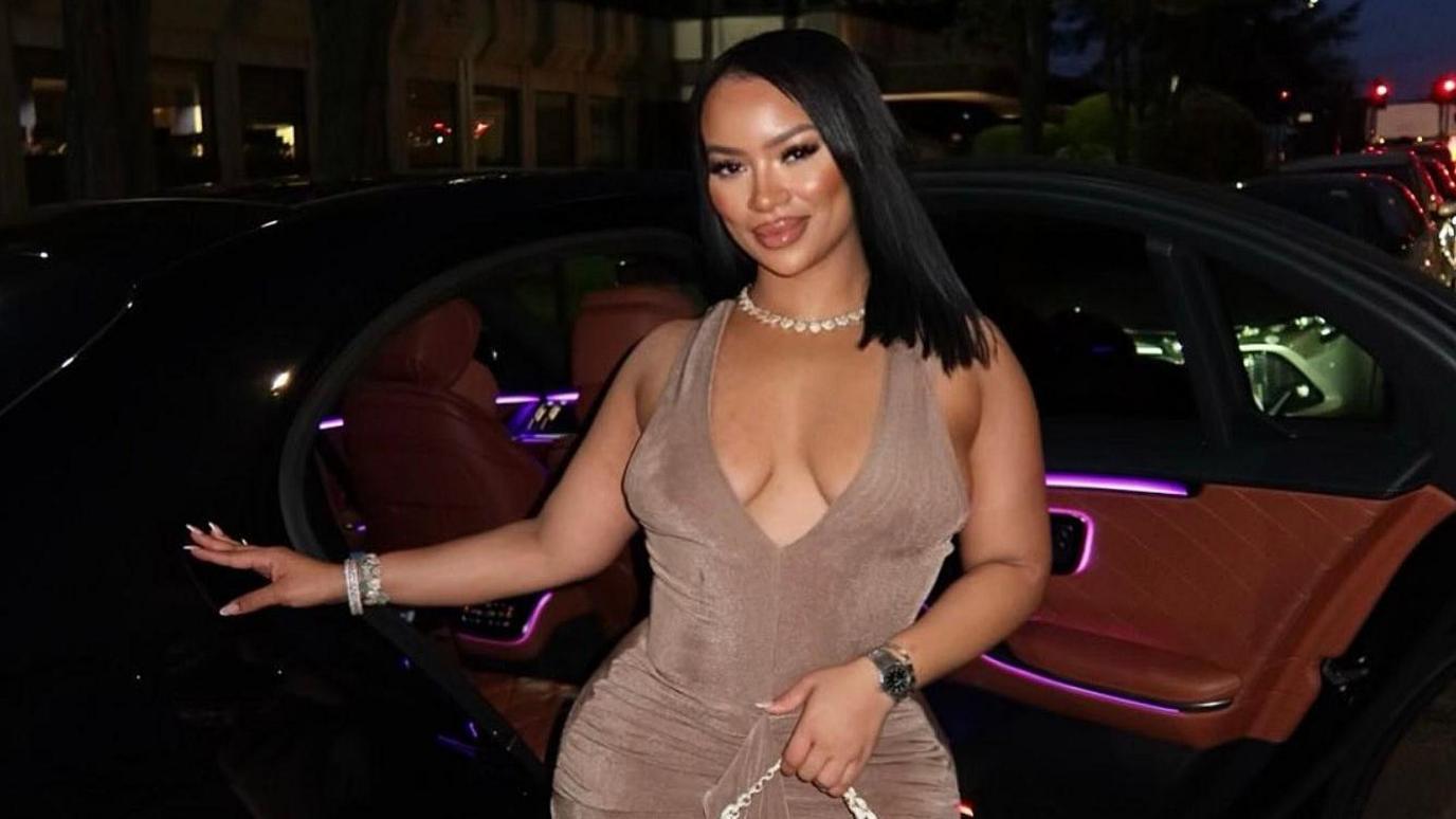 A woman in a light brown dress stands in front of a car, with a shell necklace and straight black hair.