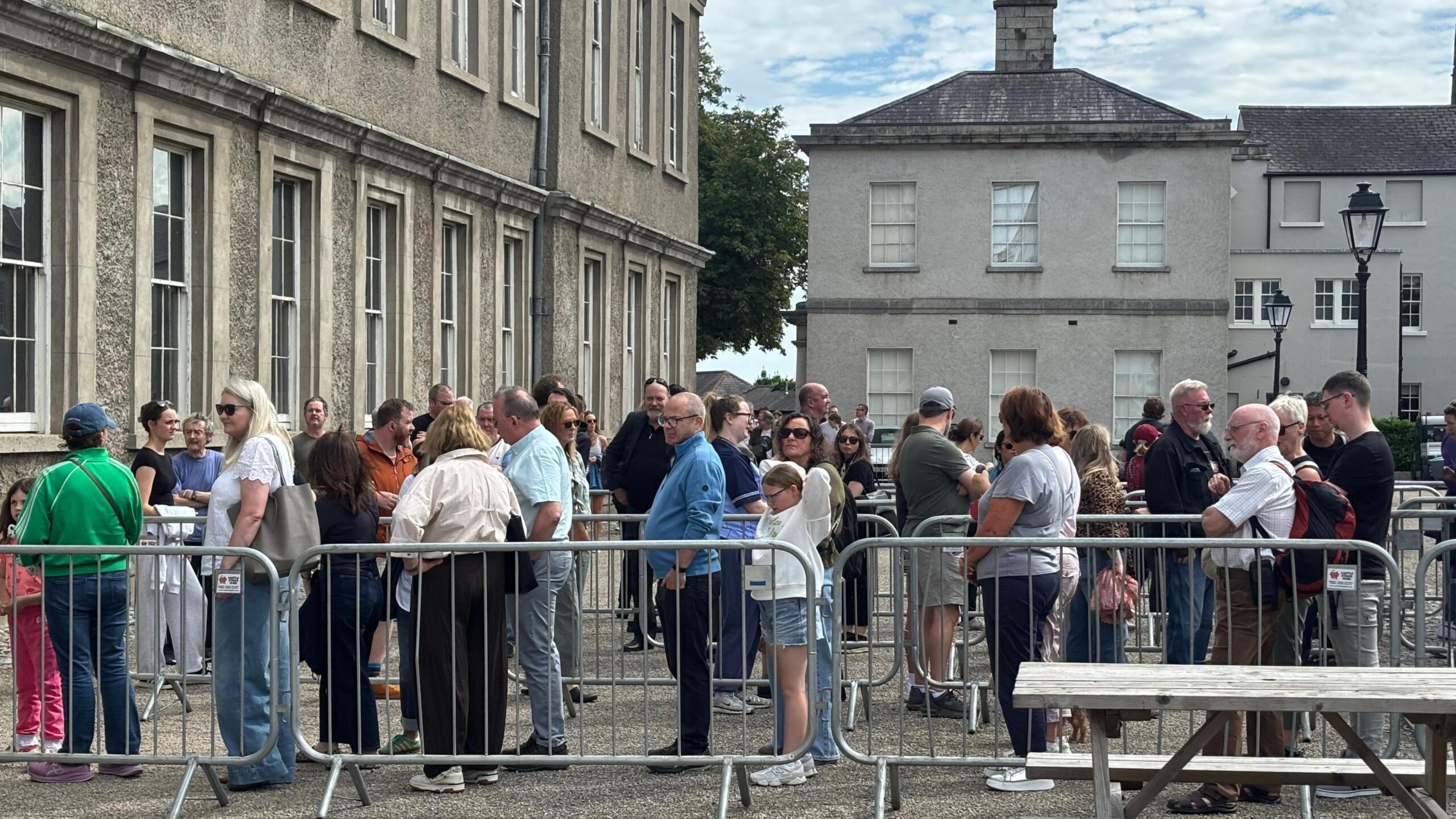 People queue in a line behind metal barriers, there is grey buildings