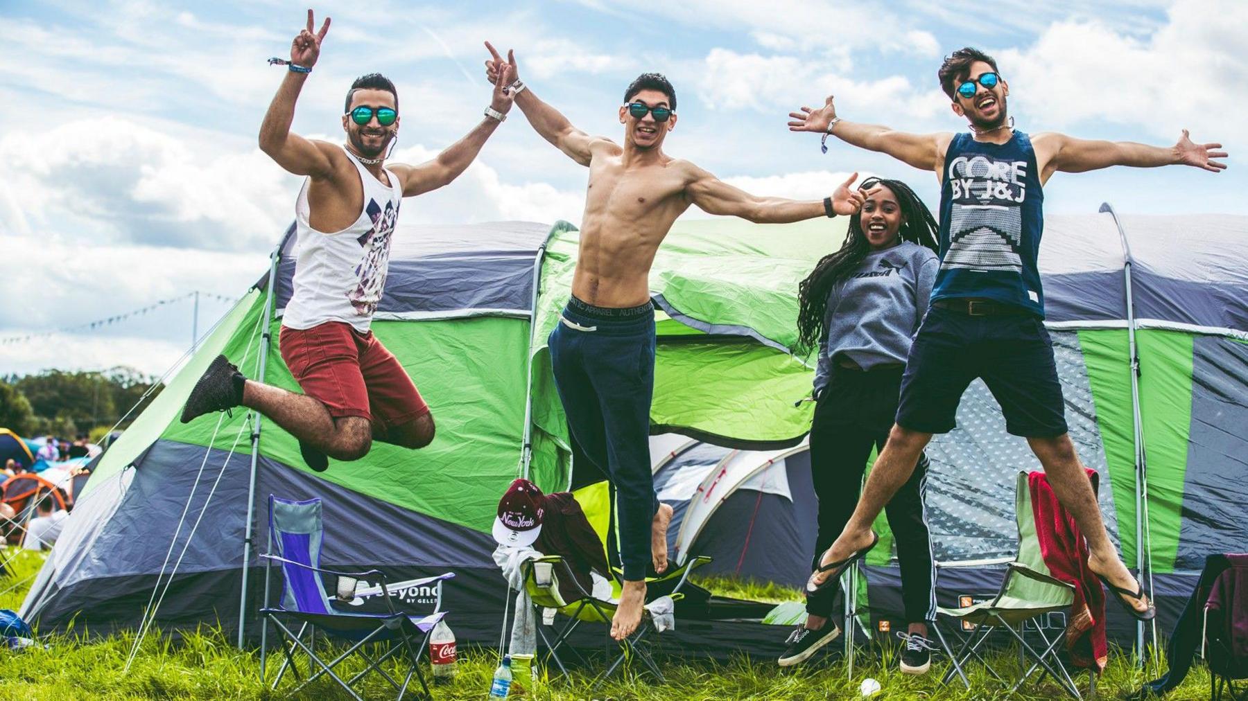 Three men and one woman are jumping and smiling at the camera outside a tent at the camping site at Creamfields.