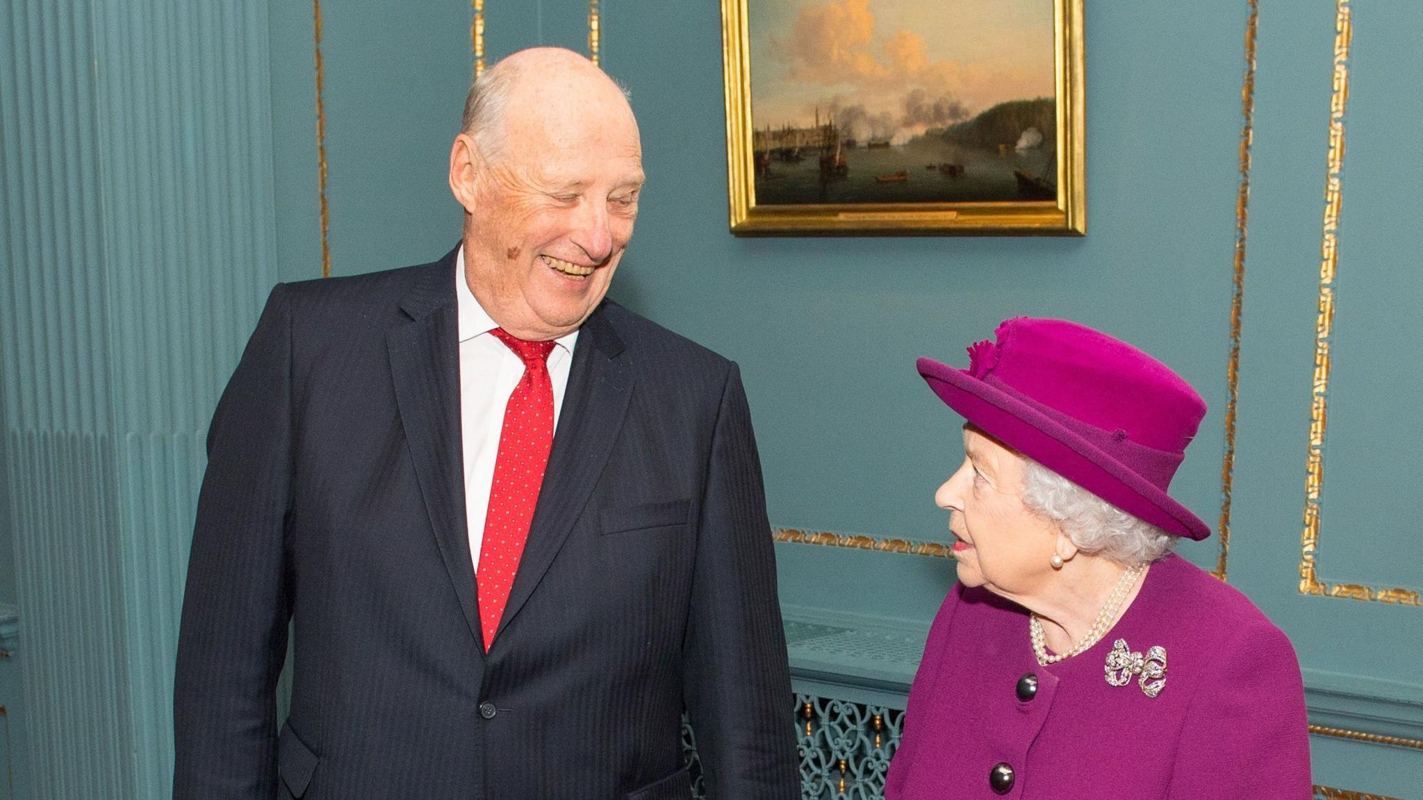King Harald wearing a suit and tie, smiles as he looks down at Queen Elizabeth II, wearing a pink suit and hat.