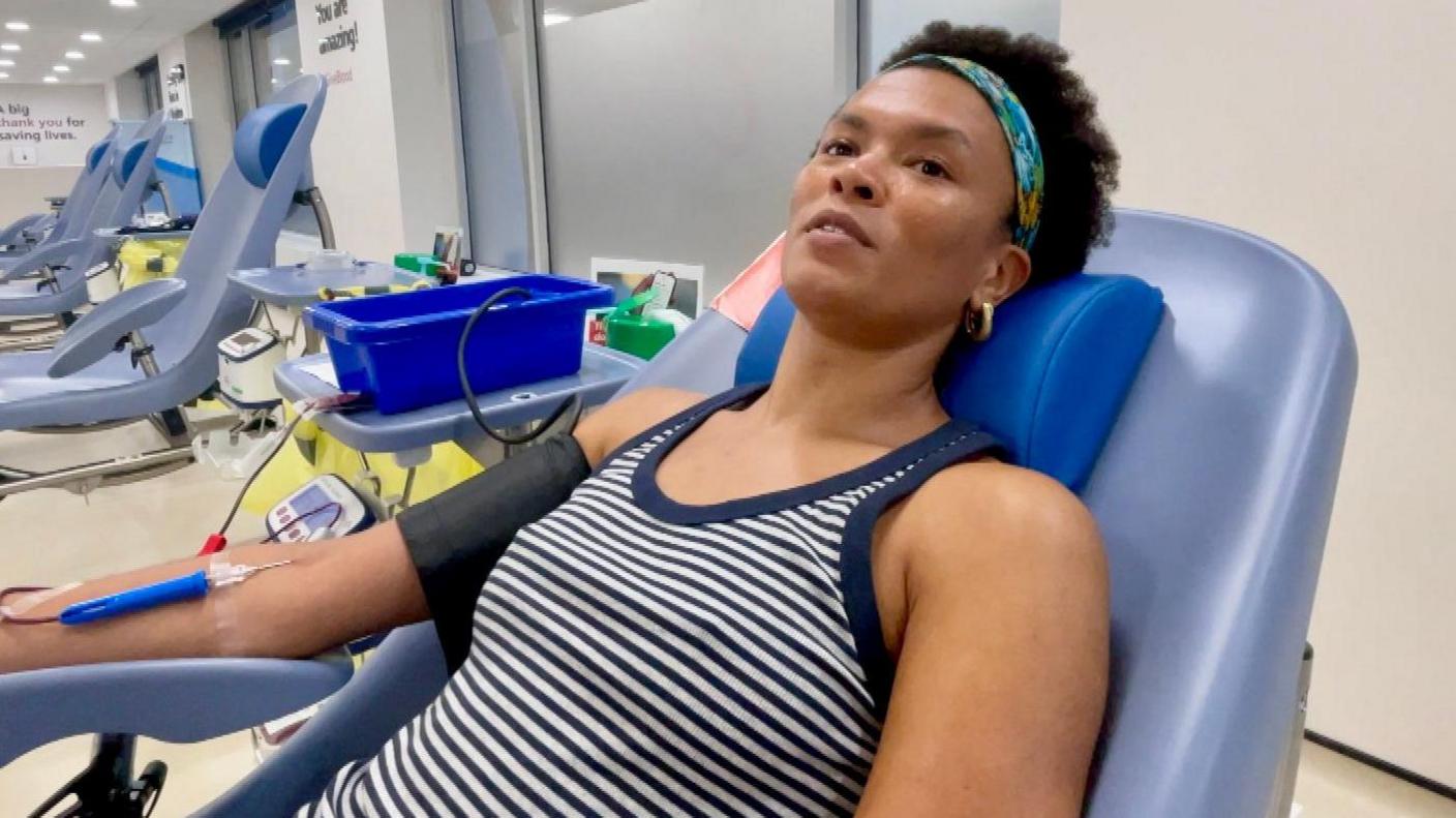 A woman lying on a clinical bed wearing a blue striped T shirt with a drip attached to her arm donating blood. There are a number of other clinical beds in the background.