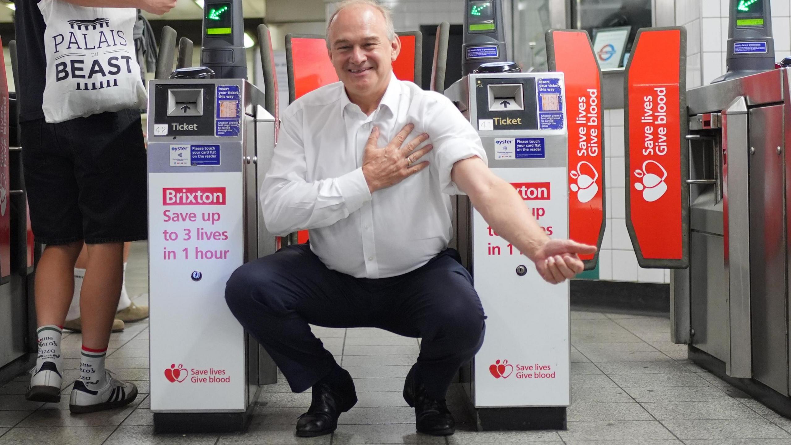 Ed Davey crouches in front of tube barriers, which are advertising the nearby blood donation centre. The adverts read: "Brixton: Save up to 3 lives in 1 hour." Ed Davey is wearing dark trousers and a white shirt with one sleeve rolled up as if about to donate blood.