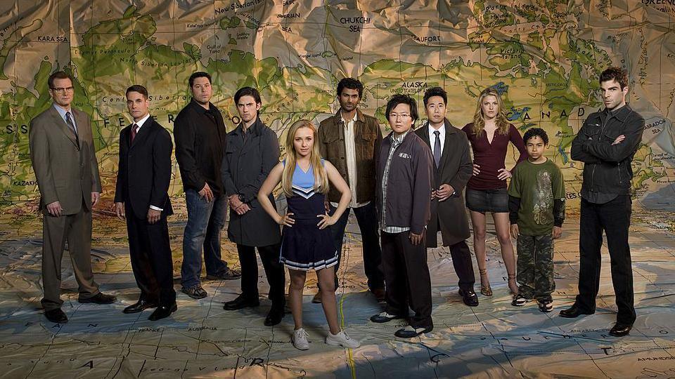 Panettiere, with straight long blonde hair and a neutral expression, wearing a blue and navy cheerleader's kit, is seen stood in the centre of the season 2 cast of Heroes in a promotional picture for the show.