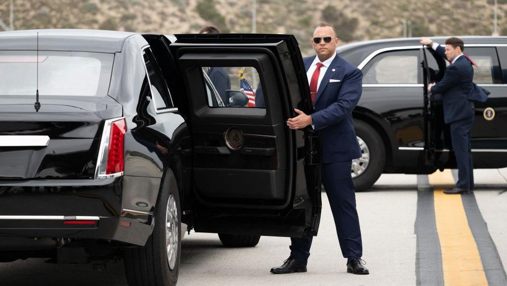 Two US Secret Service agents, both in suits, one in sunglasses, hold open the back passenger doors on two versions of the "Beast" armoured limousine.