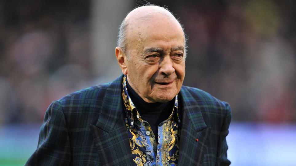 Al Fayed is wearing a suit jacket as he walks across a football pitch