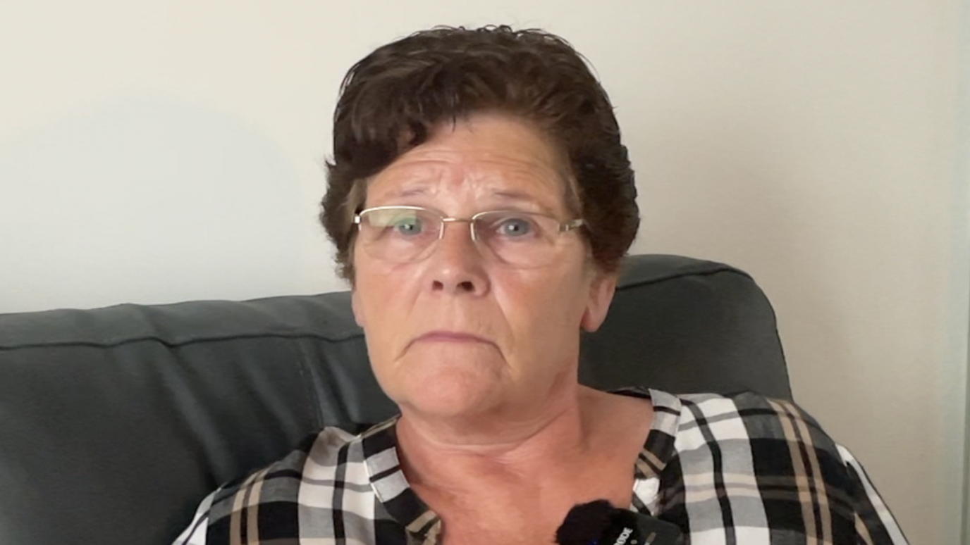 Joan is sitting on her grey sofa and looking slightly off to the right of the camera lense.  She is where a grey, white and black check shirt.  She has short dark hair, blue eyes and is wearing silver rimmed glasses.