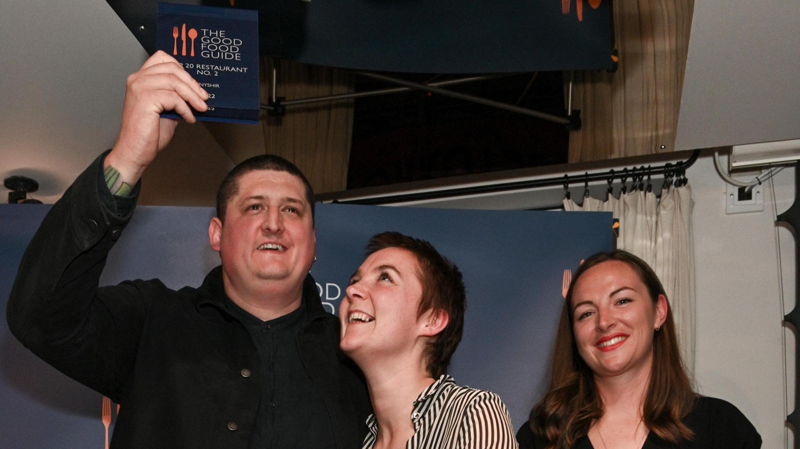 Chef Gareth Ward stands on a small stage in front of a “The Good Food Guide” backdrop. He raises an award plaque above his head while smiling, his guest, a woman, looks up at it, smiling, and a woman on the right smiles while holding a microphone and a card.