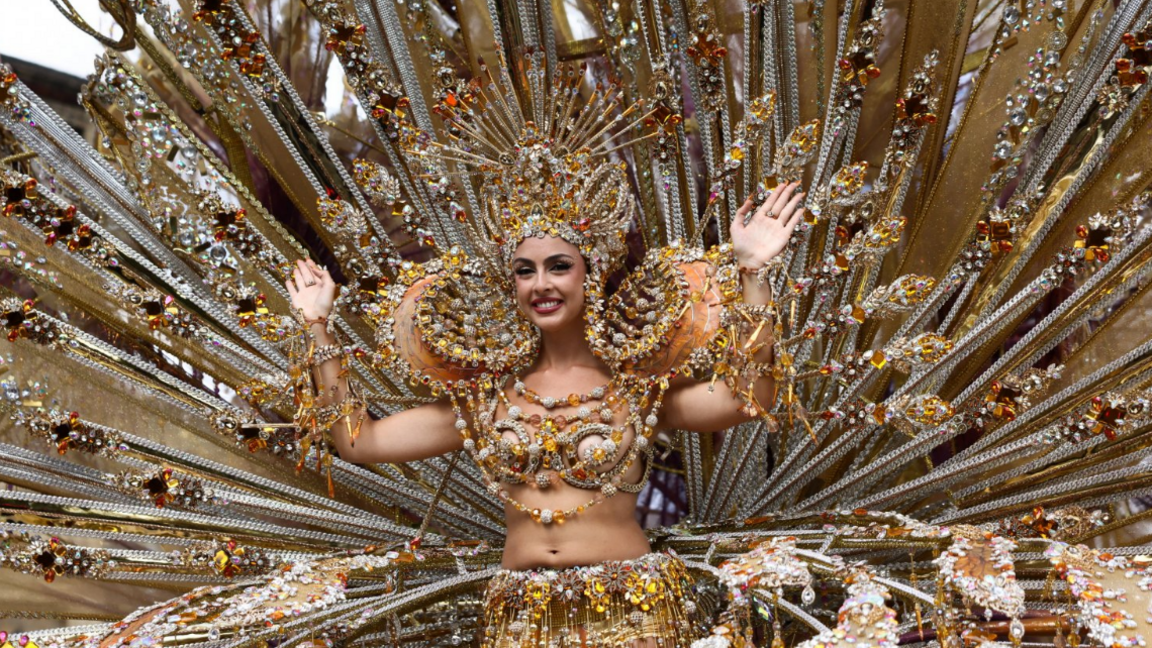 A woman in a large, fanlike gold-sequinned costume with a headdress  waves
