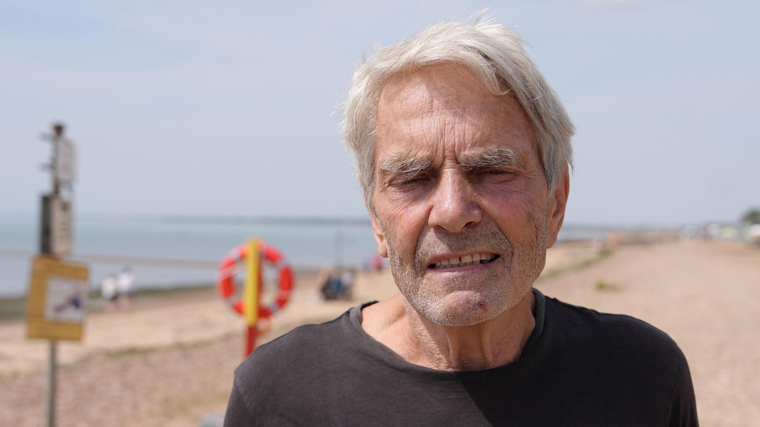 Stephen Vince has grey hair and is wearing a black top. He is standing on the beach.