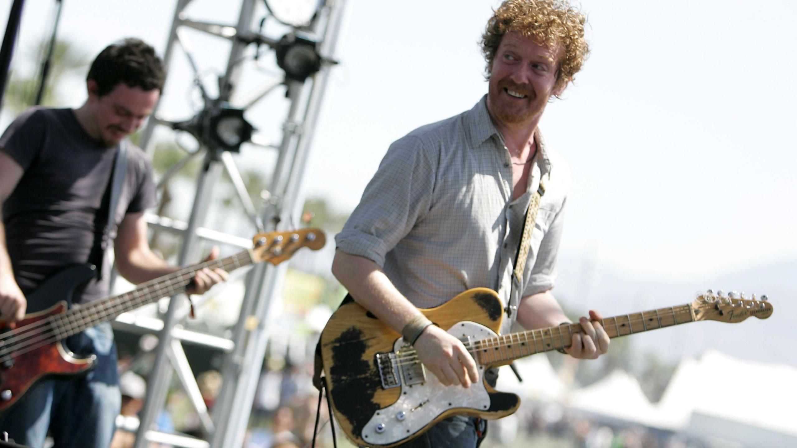 Two men on stage playing with a band, the man on the left is holding a yellow and white electric guitar and turning his head back towards a bandmate. He has an amazed smile on his face. Behind him is a man playing a bass guitar, he too is smiling while looking down at the instrument he is playing. It is daylight at a music festival.