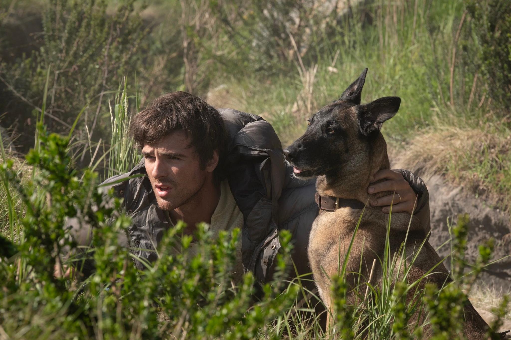 Picture of Jacob Elordi hiding in the bushes in a puffer coat with his arm around dog Jasper