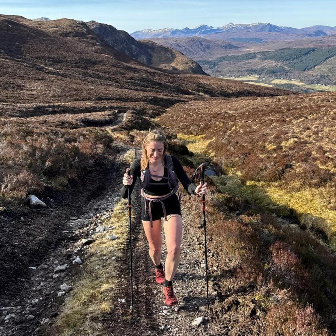 Person hiking uphill on a narrow mountain trail using trekking poles, surrounded by rolling heather-covered hills and distant snow-capped peaks under a clear blue sky.